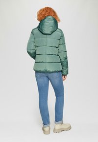 Groene pufferjack met een capuchon, met horizontale quilten en een gladde, glanzende textuur. Draag het met lichtblauwe jeans en beige laarzen.