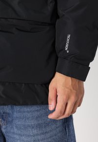Sort vandtæt jakke med en glat tekstur, der har en manchet-detail og logoet "TECH FUSION" på ærmet. Bæres over blå denim jeans.