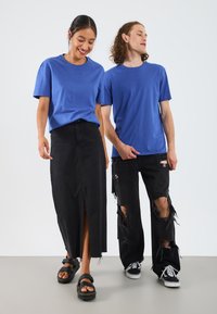 Tricouri din bumbac bleu purtate de două modele. Unul poartă o fustă din denim negru cu o deschidere; celălalt poartă blugi negri distresați și adidași.