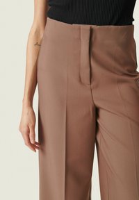 Nærbillede af en person iført brune højtaljede bukser og en sort ribbet tanktop uden ærmer, hvor der ses en del af venstre arm med en lille tatovering.