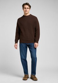 Brun ribbad stickad tröja med rund halsringning, bärs med blå jeans och bruna skor. Tröjan har långa ärmar och ett texturerat mönster.