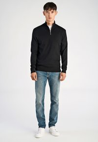 Schwarze Zip-up-Jacke mit langen Ärmeln, kombiniert mit hellblauen Jeans und weißen Sneakern, getragen von einem stehenden männlichen Model vor einem einfarbigen Hintergrund.