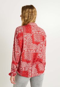 Chemise à imprimé bandana rouge avec manches longues, en tissu léger, col et poignets froncés. Motifs notables de paisley et de fleurs blancs sur l'ensemble.