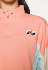 Rosa och ljusblå färgblockerad sweatshirt med halvdragkedja, hög krage och en Adidas-logotyp på bröstet. Mjuk tygstruktur.