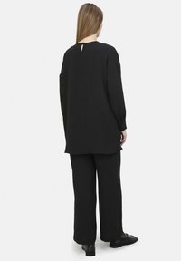 Haut noir à manches longues avec un col rond et un détail en forme de trou clef au dos, associé à un pantalon noir ample et des chaussures plates noires.