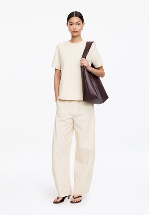Kobieta ubrana w beżowy t-shirt i spodnie, trzymająca dużą ciemnobrązową skórzaną torbę na ramię, stojąca na białym tle.