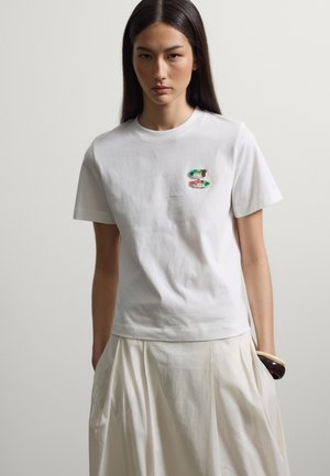 Frau mit langen dunklen Haaren, die ein weißes T-Shirt mit zwei kleinen bunten Fischen auf der linken Brust und einen cremefarbenen plissee Rock trägt.