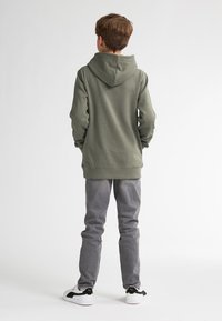 Sudadera verde con un acabado texturizado, con una gran capucha, llevada sobre pantalones grises, combinada con zapatillas negras y blancas.