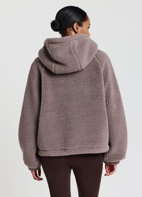 Giacca in pile oversize con cappuccio di colore taupe, caratterizzata da una superficie testurizzata e morbida. Presenta spalle scese e un orlo che si posiziona sopra la vita.