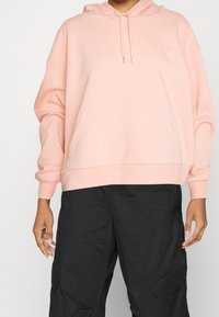 Felpa rosa chiaro realizzata in morbido cotone misto, con tasca frontale, polsini a coste e cappuccio regolabile con cordoncino. Indossata con pantaloni cargo neri.