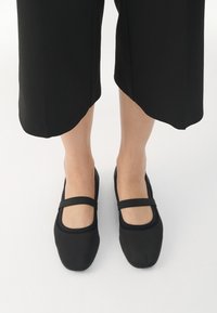 Scarpe nere flat realizzate in materiale liscio, con punta arrotondata e due strisce superiori. Indossate con pantaloni neri a tre quarti, visibile una leggera texture.