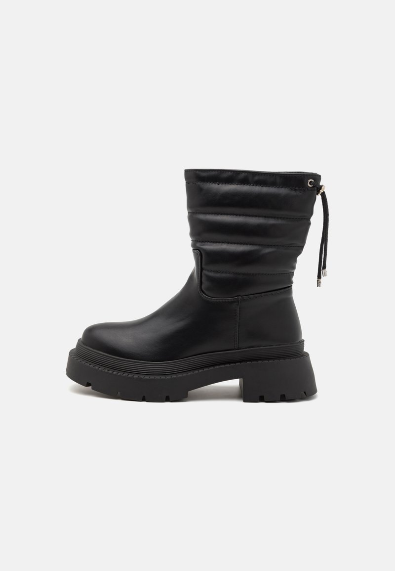 Bota de piel negra hasta el tobillo con un eje caído, suela de goma gruesa y acabado texturizado. Presenta un detalle de cordones en la parte trasera.