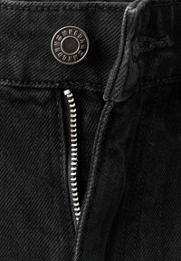 Prim-plan al unei perechi de blugi din denim negru cu fermoar argintiu parțial desfăcut și nasture metalic gravat cu textul „MNGDN”.