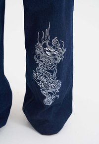 Ciemnoniebieskie spodnie z białym haftowanym wzorem smoka wzdłuż zewnętrznej nogawki. Materiał wygląda na miękki z delikatnym połyskiem.