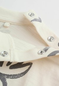 Bombažna srajca s kremno osnovo, s sivimi grafičnimi vzorci in kovinskimi zaponkami na izrezu. Gladek otip, priložnostna zasnova.