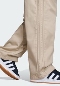Beżowe spodnie o prostym kroju, z trzema pionowymi paskami po bokach, zestawione z granatowymi trampkami z białymi paskami i gumowymi podeszwami.