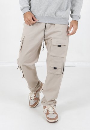 Uomo che indossa pantaloni cargo beige con più tasche e sneakers rosa e bianche, in piedi su un pavimento bianco con sfondo bianco.