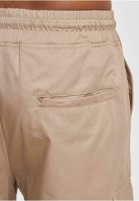 Pantaloni bej cu un band elastic la talie, având un buzunar pe partea din spate și o textură de țesătură netedă, ușor lucioasă.