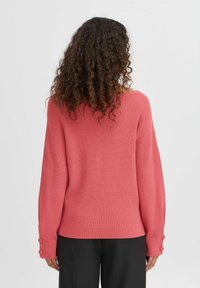 Koralni pleteni pulover z udobnim krojem, rebrasto teksturo in gumbnimi detajli na manšetah. Zadnji pogled prikazuje dolge rokave in preprost dizajn.