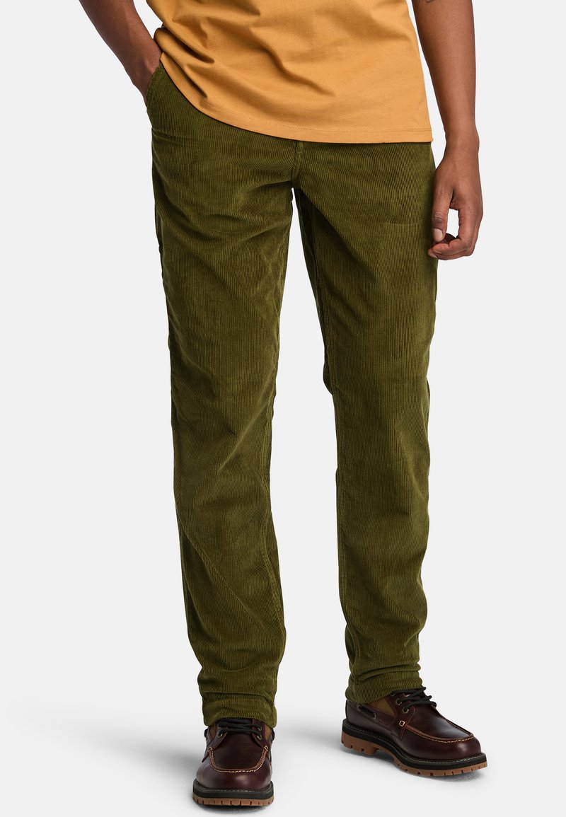 Uomo che indossa pantaloni in velluto a coste verde oliva, una camicia giallo senape e scarpe in pelle marrone scuro, in piedi con le mani rilassate.