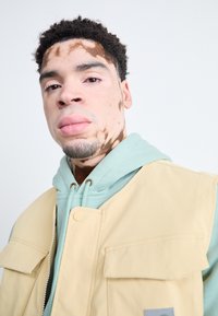 Beige Weste mit vorderen Taschen, die über einem hellgrünen Hoodie getragen wird. Glattes Material mit kontrastierenden Texturen, das einen grauen Akzent auf der Weste aufweist.