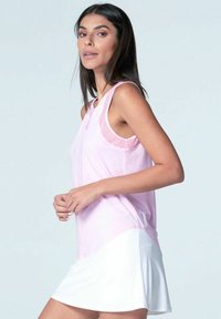 Mujer con cabello largo y oscuro, vistiendo una blusa sin mangas de color rosa claro y una falda blanca, de pie frente a un fondo claro y sencillo.
