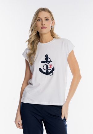 Blondynka w białym bezrękawniku z granatowym nadrukiem kotwicy i czerwonymi inicjałami, w granatowych spodniach, stojąca na białym tle.