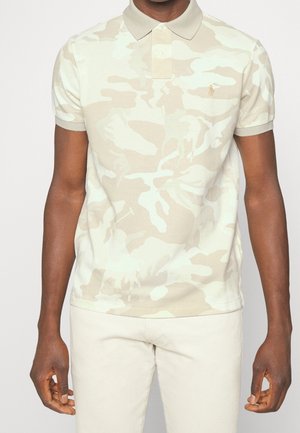 Mężczyzna ubrany w beżowo-białą koszulkę polo we wzór kamuflażu z kołnierzykiem oraz jasnobeżowe spodnie, ramiona swobodnie opuszczone wzdłuż ciała.