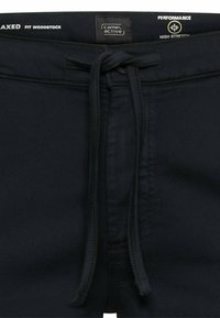 Black high-stretch Camel Active pants with drawstring waist, labeled "Relaxed Fit Woodstock" and "Performance High Stretch."