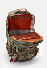 Plecak w kamuflażowym wzorze z wieloma zamkami błyskawicznymi, beżowymi paskami, otwartą główną komorą pokazującą jasnopomarańczowe i siatkowe kieszenie wewnętrzne.