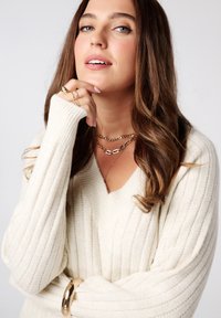 Crème gebreide trui met ribbeltextuur, v-hals en aansluitende boorden. Gouden sieraden, waaronder gelaagde kettingen en ringen, completeren de look.