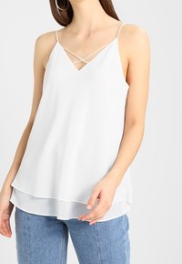 Valkoinen, hihaton pusero, jossa on v-muotoinen kaula-aukko ja ristikkäin oleva hihna. Ominaisuuksiin kuuluu kerroksellinen helma ja kevyt kangas, joka antaa pehmeän ilmeen.