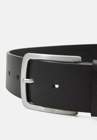 Curea neagră din piele cu o cataramă dreptunghiulară din metal argintiu, gravată cu marca. Textură netedă, cu mai multe găuri de ajustare de-a lungul curelei.