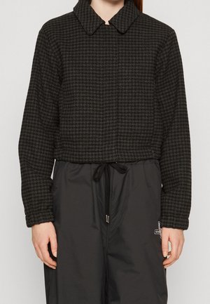 Vrouw draagt een donker houndstooth-patroon cropped jas met lange mouwen en zwarte broek met trekkoord.