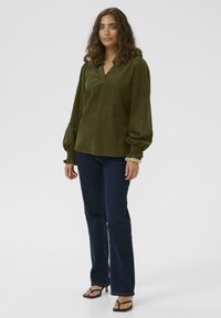 Ženska s kodrasto lasmi, oblečena v olivno zeleno dolgo majico s dolgimi rokavi, temno modre kavbojke in črne sandale s petami, stoji na beli ozadju.