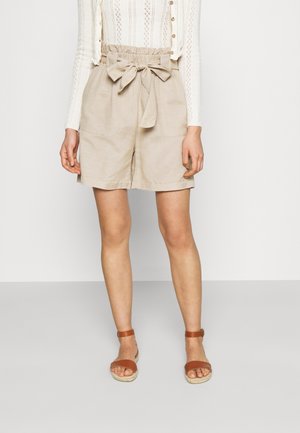 Donna che indossa pantaloncini beige a vita paperbag con cintura annodata e sandali con cinturino alla caviglia color cuoio, in piedi su sfondo semplice.