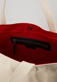 Bolsa de lona con un exterior crema y un interior rojo. Presenta un bolsillo con cremallera negro en el interior, costuras reforzadas y asas resistentes.