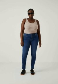 Prugasta majica bez rukava u svijetlosmešavoj narandžastoj, plavoj i bijeloj; plave uske traperice; crne cipele; sunčane naočare; stoji protiv neutralne pozadine.