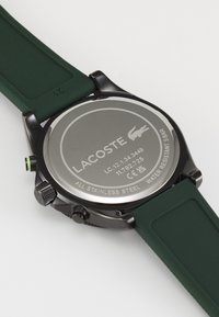 Montre en acier inoxydable noir avec un bracelet en silicone vert. Le dos présente un logo et un texte gravés, indiquant la résistance à l'eau et les détails du modèle.