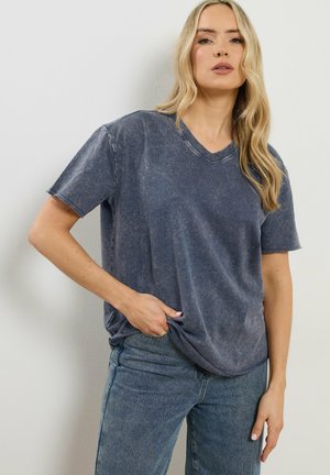 Blondynka w wyblakłym granatowym T-shircie z dekoltem w serek i niebieskich dżinsach, stojąca z jedną ręką w kieszeni na tle białej ściany.