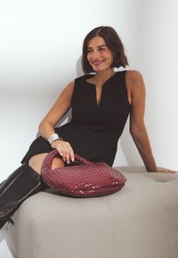 Burgundská tkaná kabelka s lesklým povrchom, v kombinácii s čiernymi bezrukávovými šatami. Sedí na svetlosivej podnožke; viditeľné čierne čižmy po kolená.