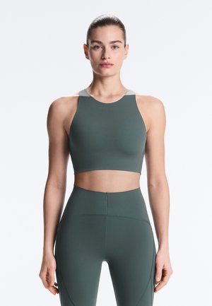 Žena v tmavozelenom športovom crop tope s vysokým golierom a zodpovedajúcich legínach s vysokým pásom, stojaca čelom dopredu na bielom pozadí.
