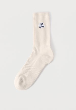 Enkelsok in roomkleur met ribbelstructuur en een klein blauw geborduurd ontwerp vlak bij de bovenkant, op een witte achtergrond.