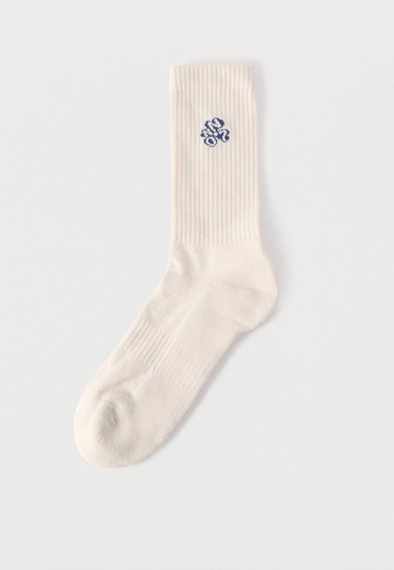 Μονή ριγωτή κάλτσα σε κρεμ χρώμα με μικρό μπλε κεντητό σχέδιο κοντά στην κορυφή, σε λευκό φόντο.