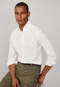 Camisa blanca de botones con mangas arremangadas, que presenta un pequeño logotipo azul. Combinada con pantalones verde oliva y un cinturón negro. Diseño sencillo, material de algodón.