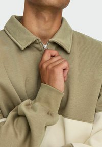 Collared sweatshirt in muted olive green with a half-zip closure. Features ribbed cuffs and a cream accent along the lower hem. Soft texture.