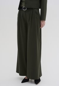 Grüne weit geschnittene Hosen aus glattem Stoff, mit Falten und hohem Bund. Getragen mit einem schwarzen Gürtel und spitzen schwarzen Schuhen.