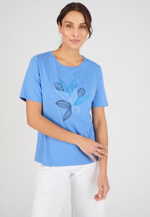 Woman in blue short-sleeve shirt with blue and black leaf design, white pants, standing against plain light background, smiling gently.