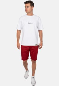 Witte t-shirt met korte mouwen en zwarte tekst, gecombineerd met rode opgerolde short en witte sneakers. Eenvoudig, casual outfitontwerp.