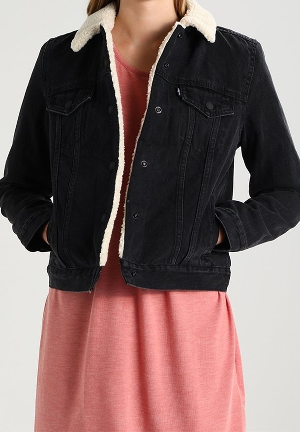 Veste en denim noir avec un col en fausse fourrure crème, dotée de fermetures à boutons, deux poches poitrine et une coupe courte, portée sur une robe rose.